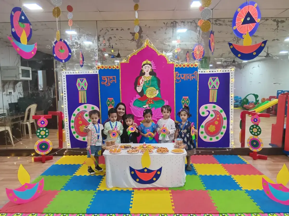 Children holding colourful crafts in front of a festive display with paper diyas and a sweets table